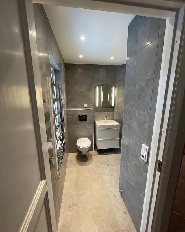 Badrum med grå marmor, handfat, toalett och handdukstork. Dörröppning i förgrunden